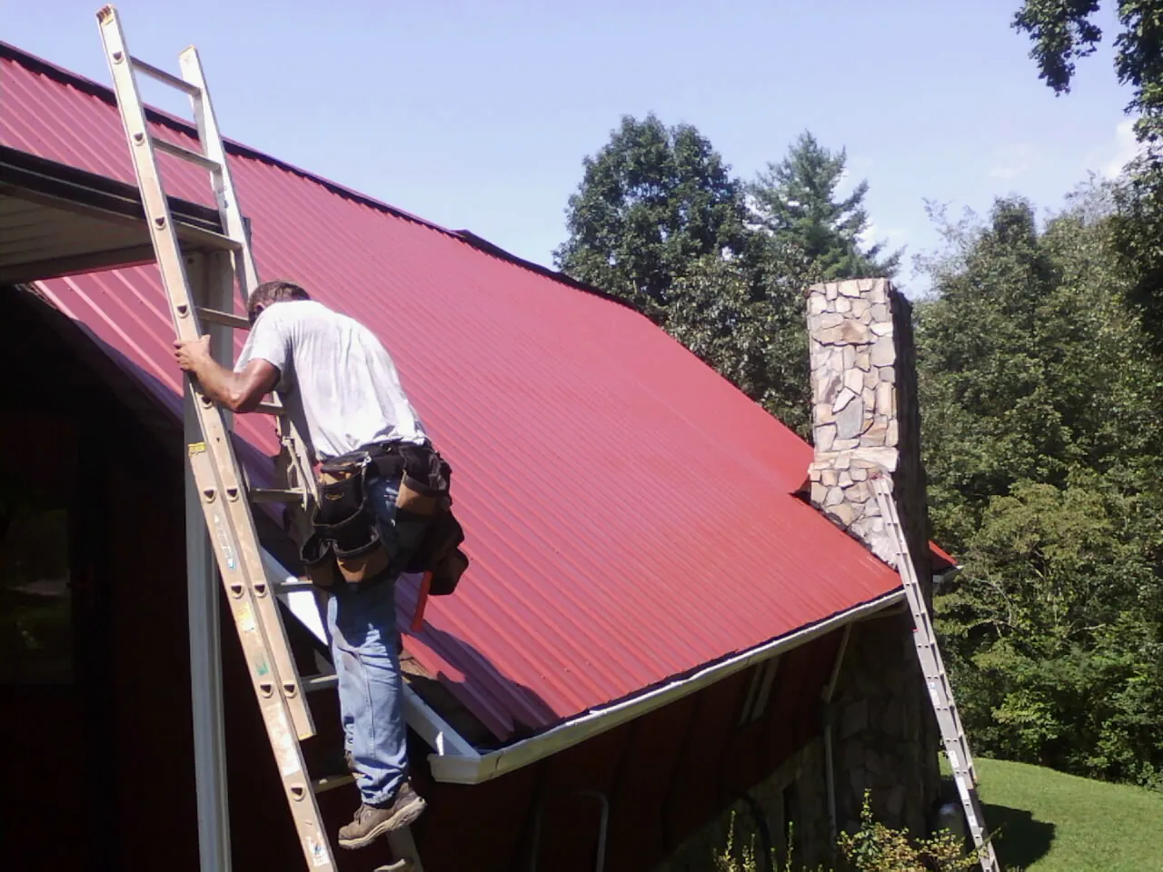 Roofer on a ladder beside a red metal roof and stone chimney — Soffit & Fascia Repair by Roofing Brothers in Lewisport
